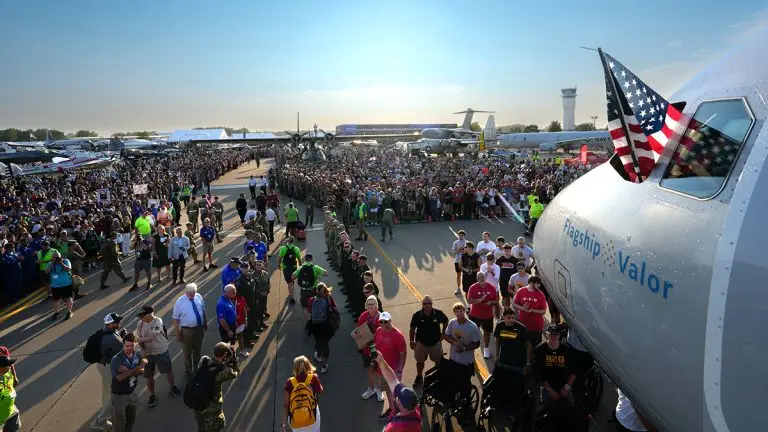 Crowd gathers on an airstrip around a large military aircraft, with an American flag in the cockpit and the plane marked 'Flagship Valor'.
