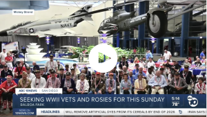 Group photo of WWII veterans seated together at a museum exhibit with fighter jets overhead, news banner at bottom reading 'SEEKING WWII VETS AND ROSIES FOR THIS SUNDAY'