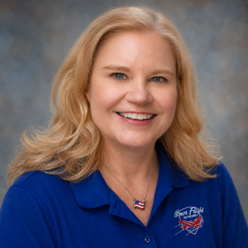 Smiling blonde woman in a blue polo with Honor Flight Network logo, posing for a portrait portrait against a neutral backdrop.